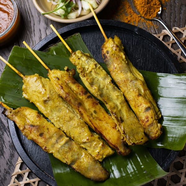 Six chicken satay skewers on banana leaves, with peanut sauce and cucumber salad.