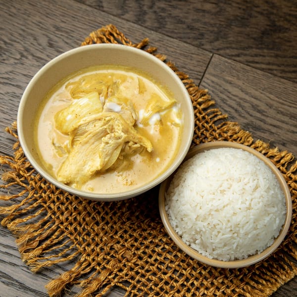 A bowl of yellow curry with chicken and a side of white rice on a woven placemat.