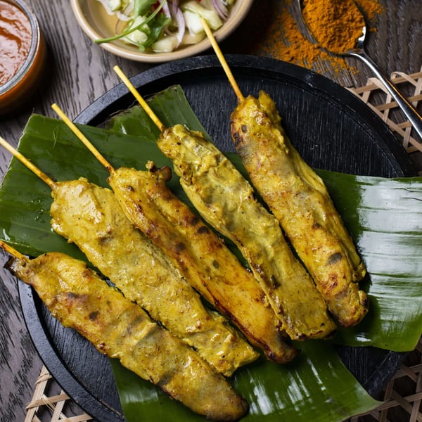 Five grilled chicken satay skewers on a banana leaf, with peanut sauce and cucumber salad.
