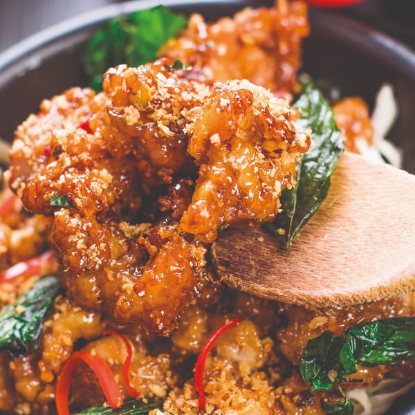 Close-up of crispy Thai chicken with sweet glaze, basil, and chili, being served with a wooden spoon.